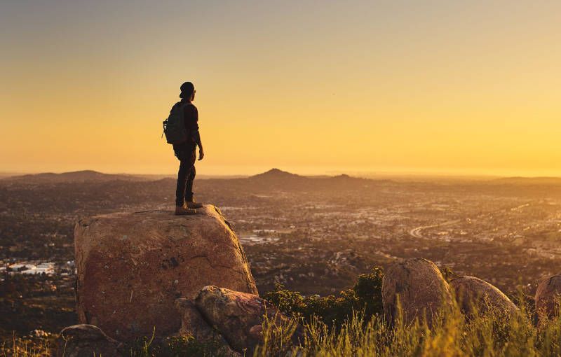 A man is standing on top of a rock overlooking a city at sunset.