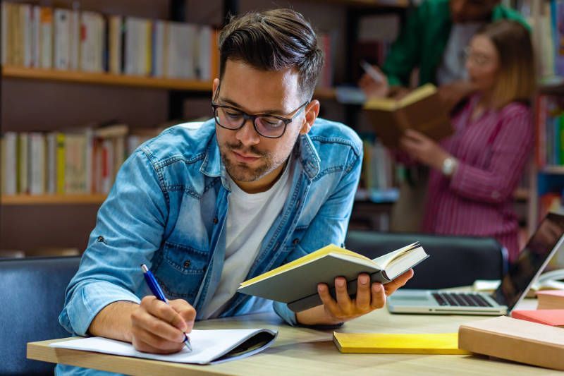 A man is sitting at a desk in a library holding a book and writing in a notebook.