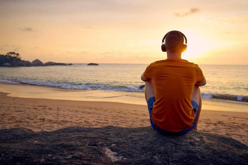 A man is sitting on a rock on the beach listening to music.