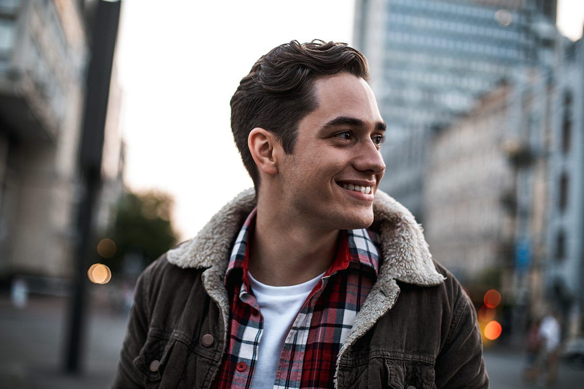 A young man wearing a plaid shirt and a jacket is standing on a city street.