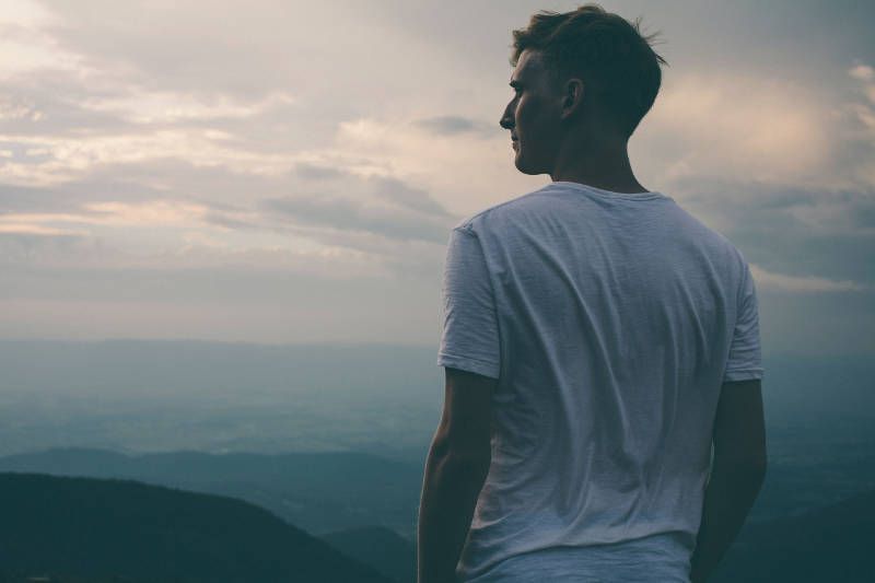 A man in a white shirt is standing on top of a mountain looking at the sky.