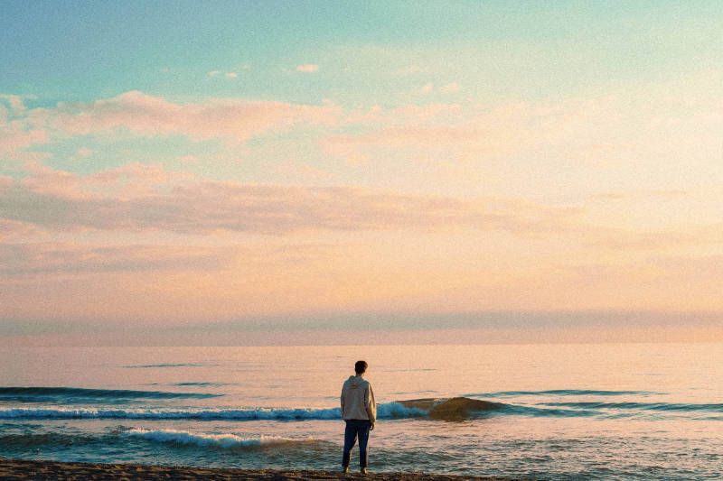 A man is standing on a beach looking at the ocean at sunset.