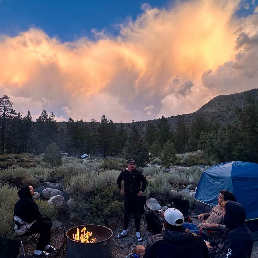 People camping around a campfire in a mountain setting at sunset.