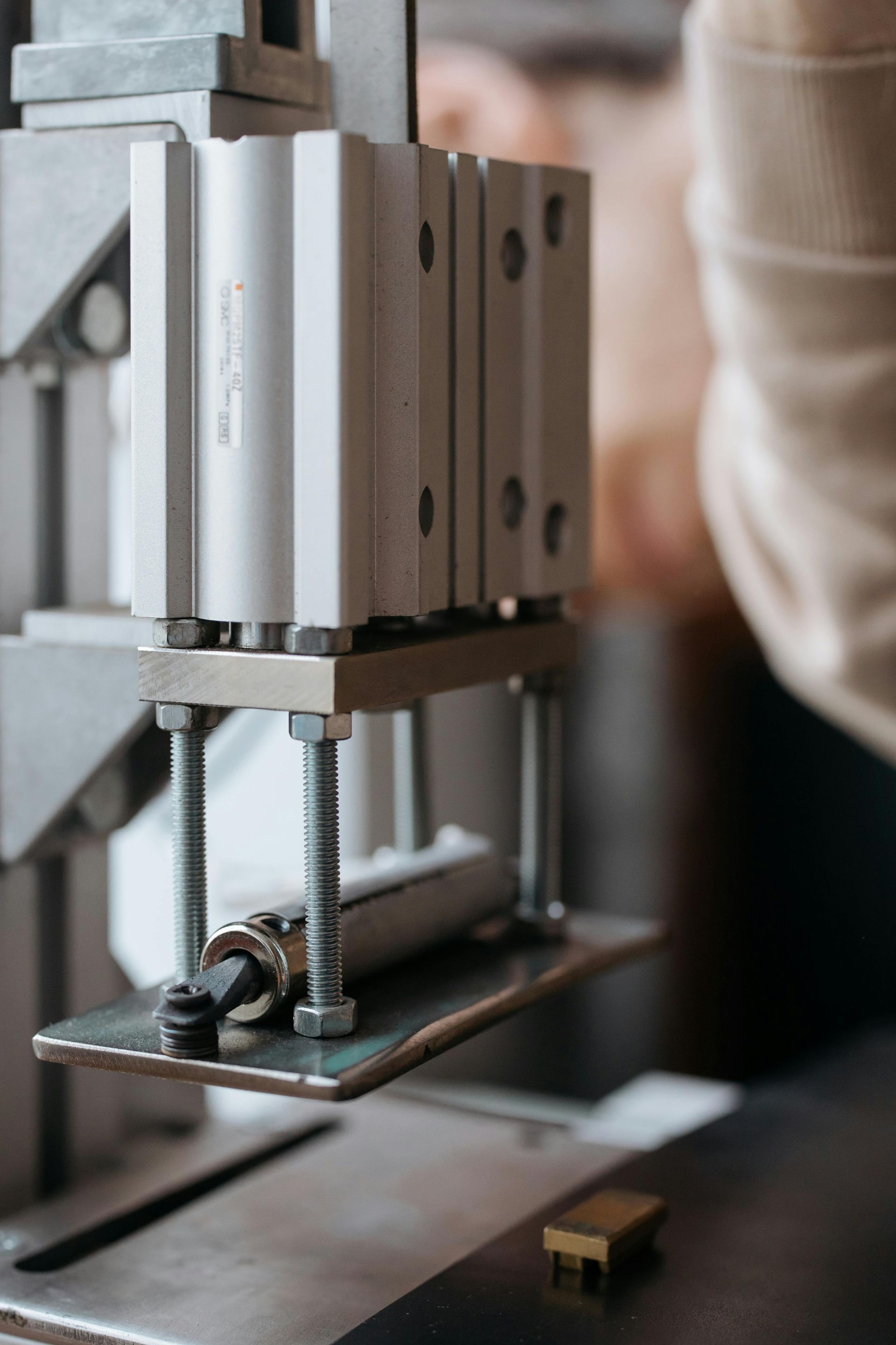 Close-up of a pneumatic press, silver cylinders on a metal plate, pressing down on a small, gold-colored block.