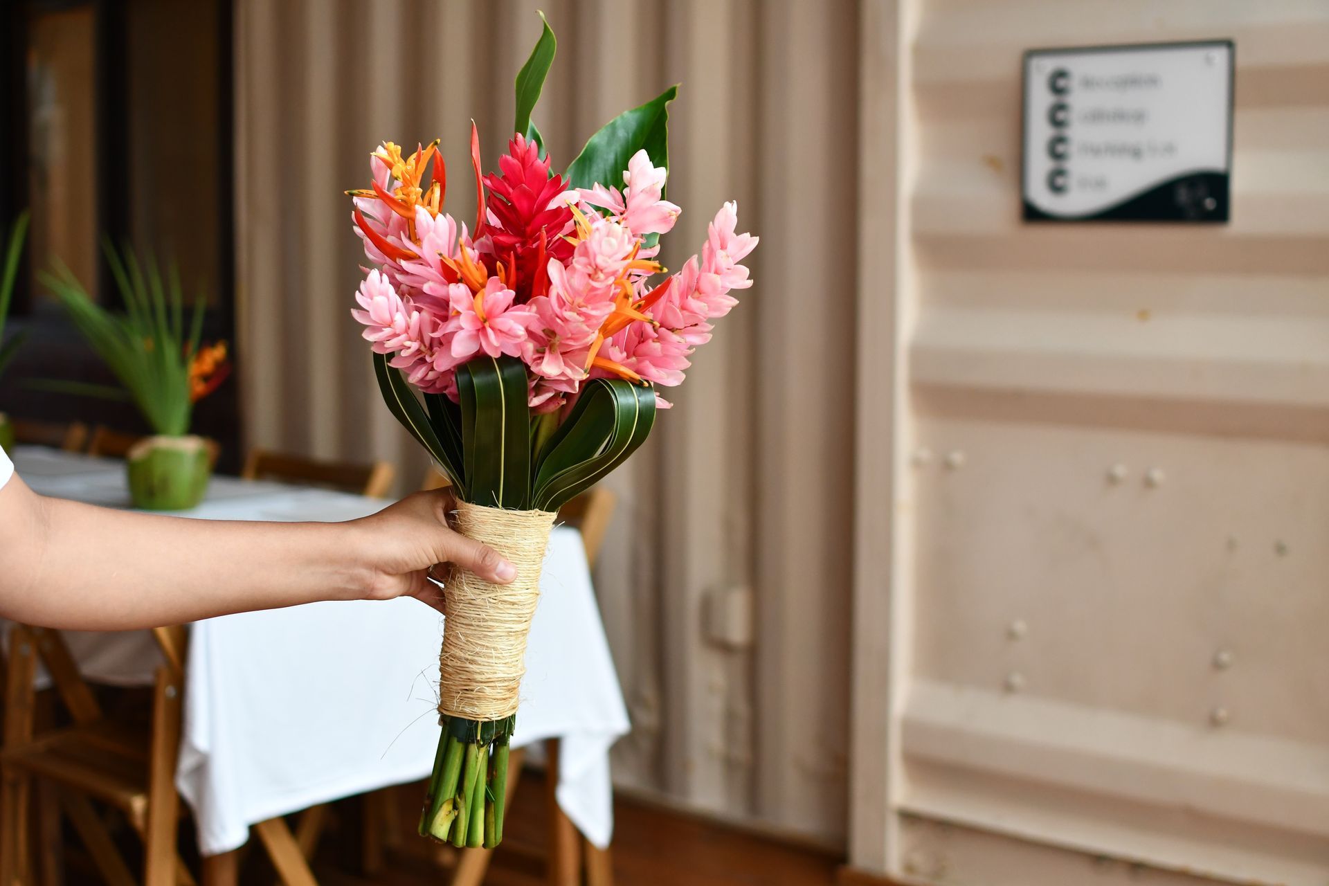 Person holding a bouquet of colorful ginger flowers, wrapped in natural twine, near a table.