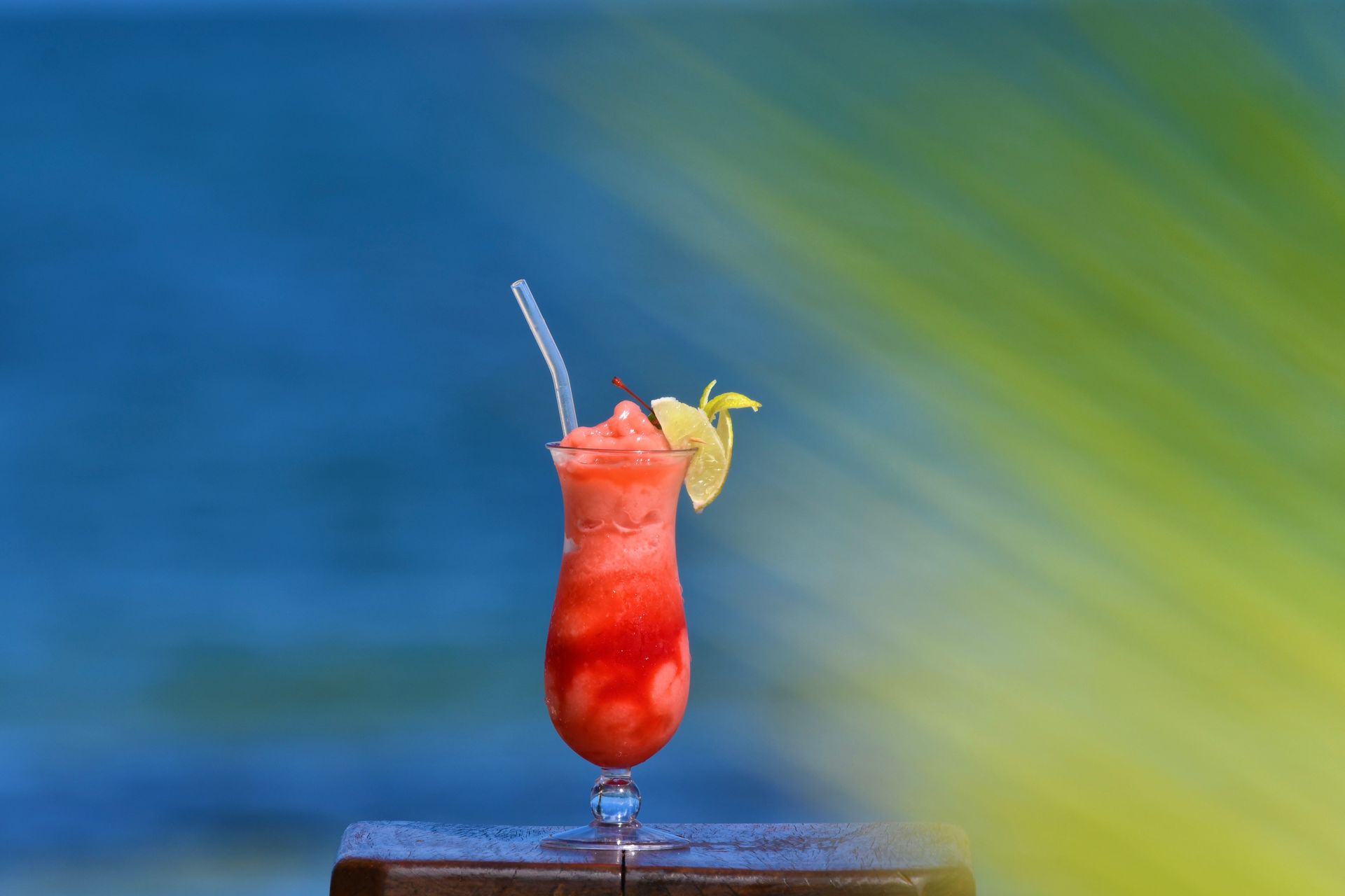 Strawberry cocktail with a straw and garnish, set against a blue ocean and green foliage.