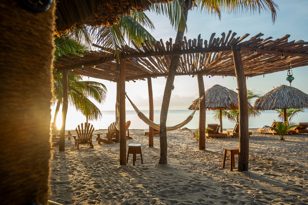 Beach scene with thatched-roof structures, hammocks, chairs, palm trees, and sunlight over the ocean.