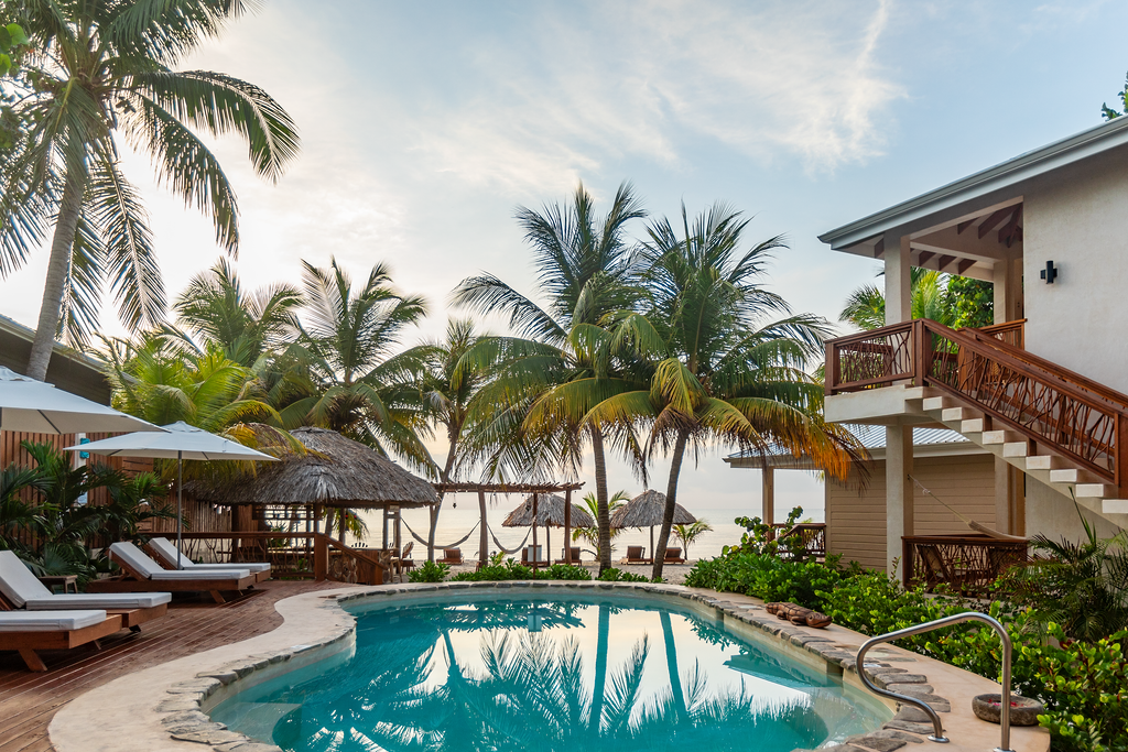 Poolside view with palm trees, beach, and white buildings; sun loungers beside the pool.