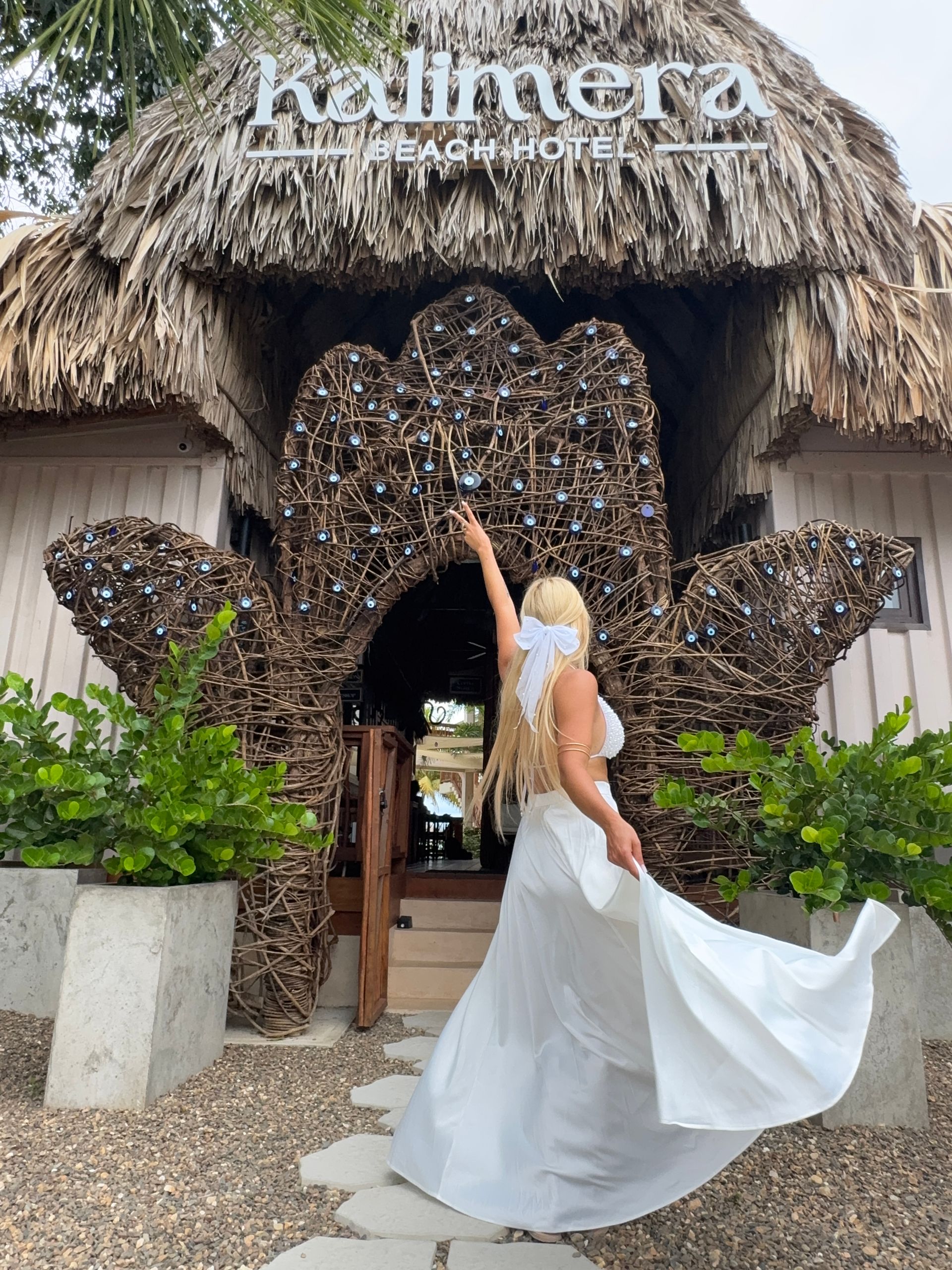Woman in white dress in front of a thatched-roof hotel entrance, raising her arm.