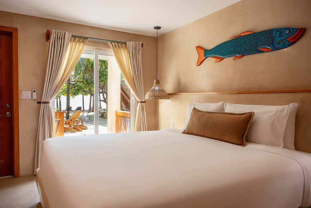 Bedroom with wooden bed, desk, artwork, and ceiling fan. Pale walls, neutral tones, natural light.