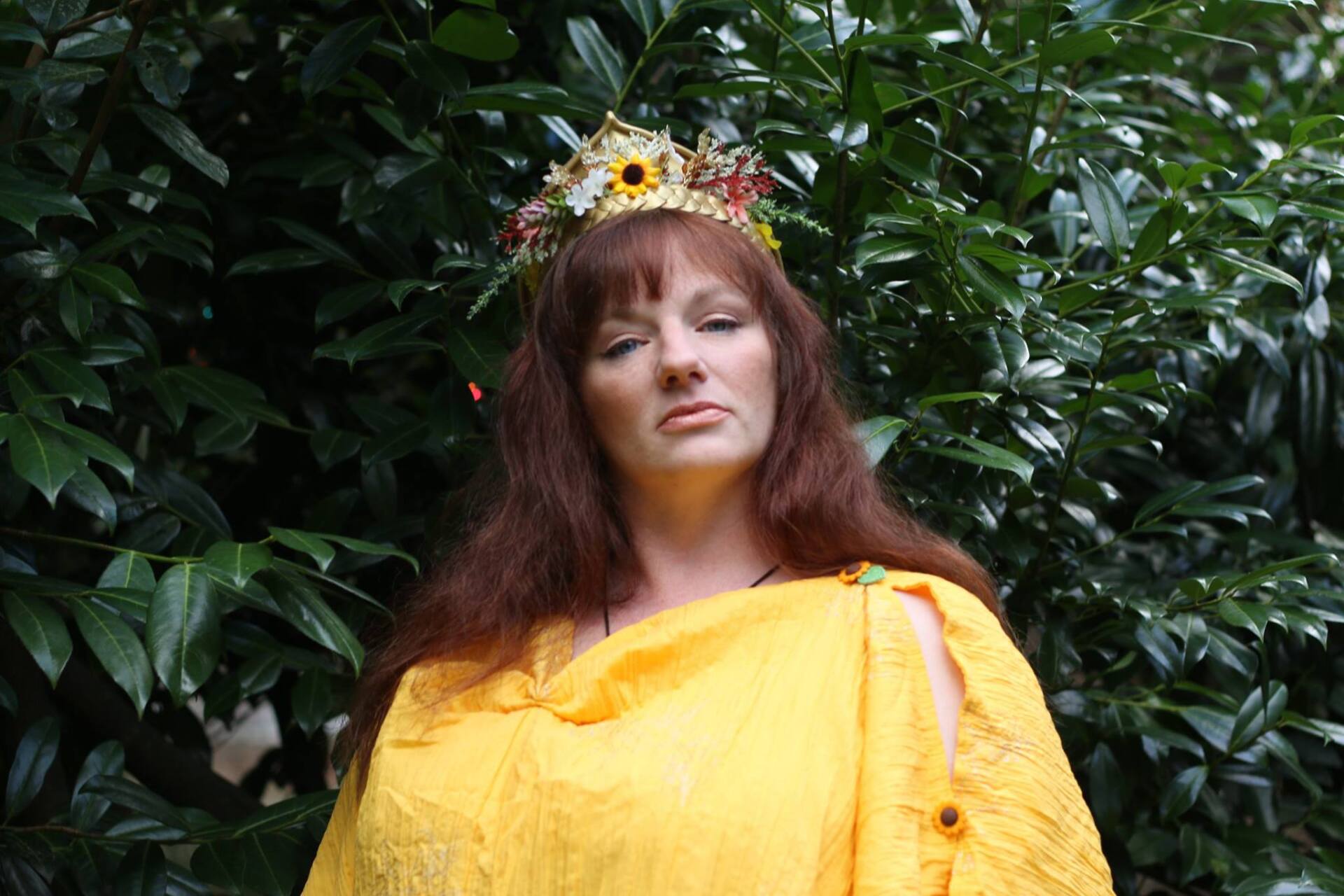A woman wearing a yellow dress and a crown is standing in front of a tree, Isidora LaVeau representing Persephone.