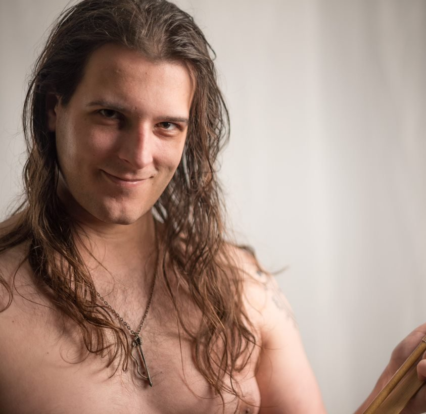 A shirtless man with long hair is smiling and holding a wooden stick, Ranger Monomachos.