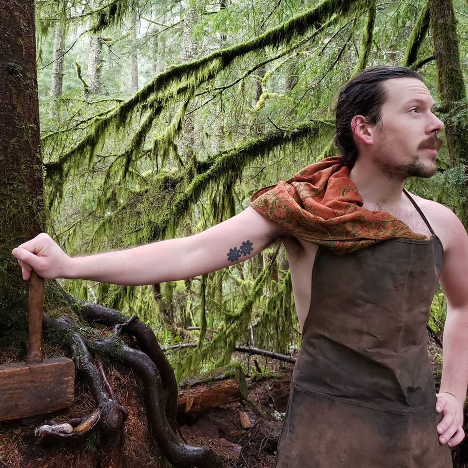 A man with a tattoo on his arm is standing in the woods, representing Hephaestus. 