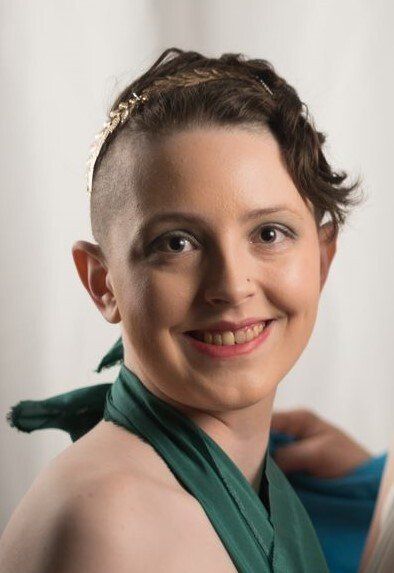 A woman with a shaved head and a green scarf around her neck, Kel representing Euphrosyne.