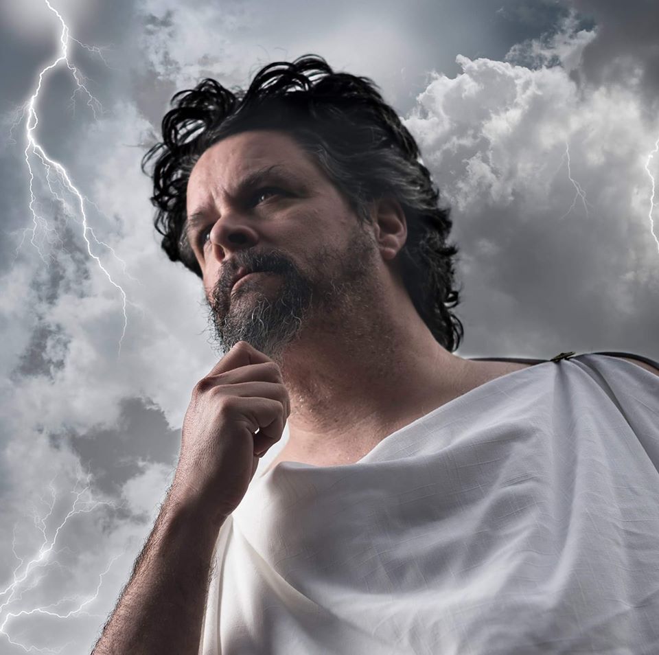 A man with a beard is standing in front of a cloudy sky with lightning, representing Zeus.