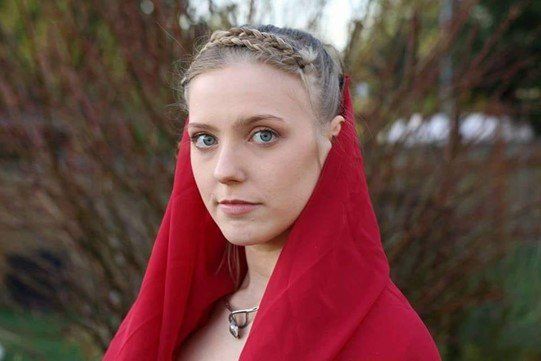 A woman wearing a red veil and a necklace is standing in front of a tree, representing Pythia.