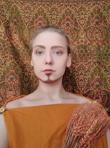 A woman with a beard is holding a bouquet of wheat in front of a wall.