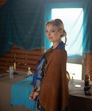 A woman in a brown poncho is standing in front of a window, representing Athena.