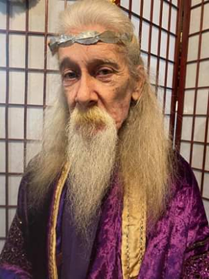 A man with long hair and a beard is wearing a purple robe and a crown, Terry Riley.