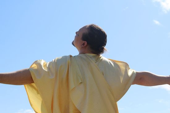 A man in a yellow robe is standing with his arms outstretched against a blue sky.