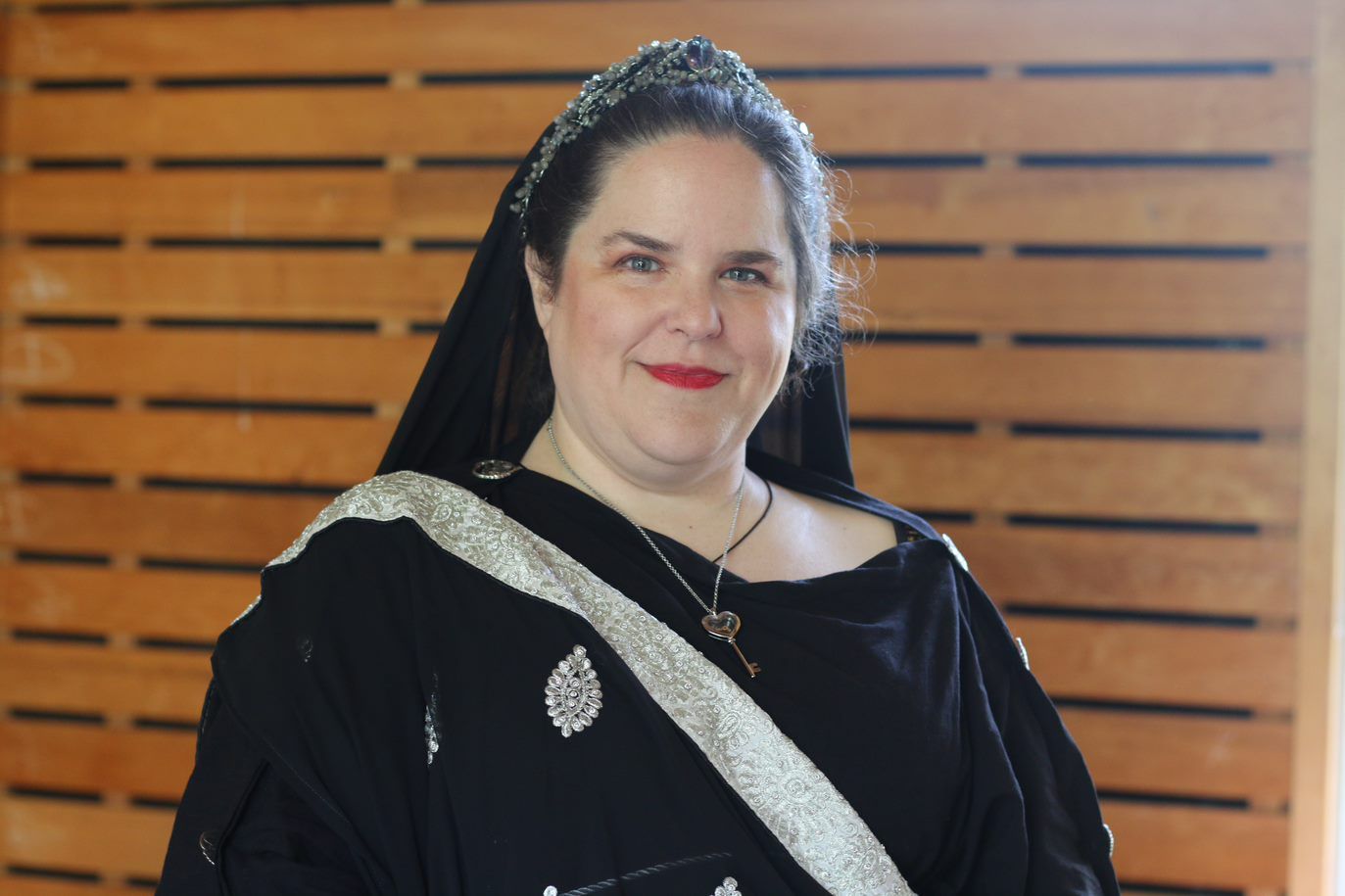 A woman wearing a black dress and a tiara is smiling in front of a wooden wall, representing Hekate.
