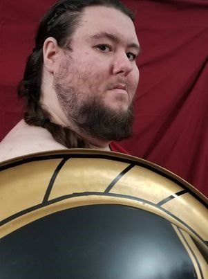 A man with a beard is holding a shield in front of a red curtain, Nikodaemus Shadowsky representing Ares.