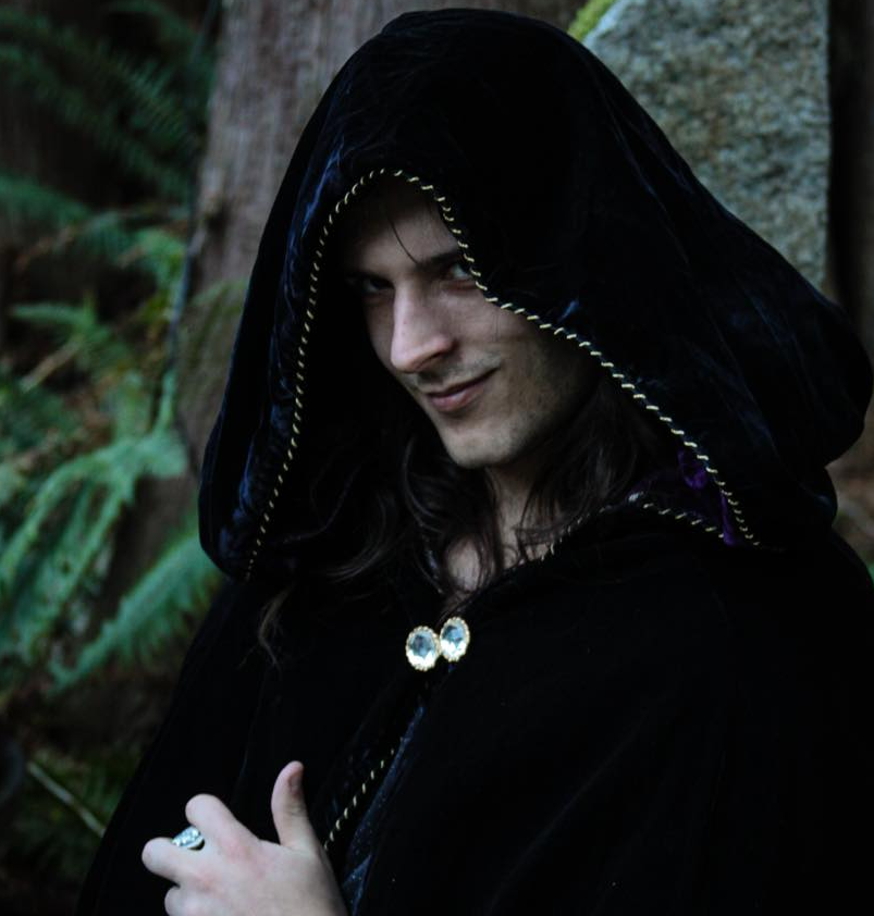 A man with long hair is wearing a black cape with a hood, representing Hades.