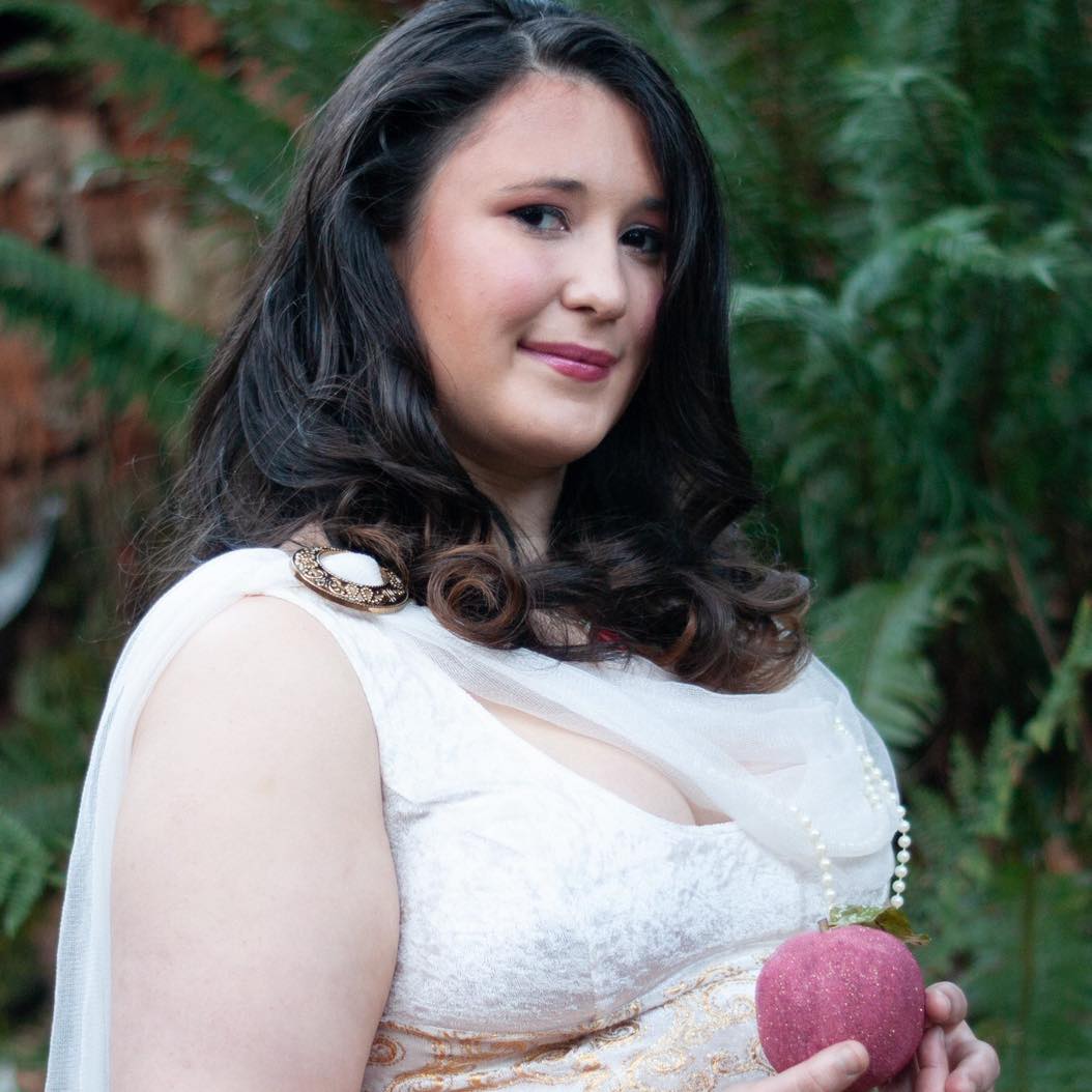 A woman in a white dress is holding a pink apple, Raven Wolfenone representing Persephone.