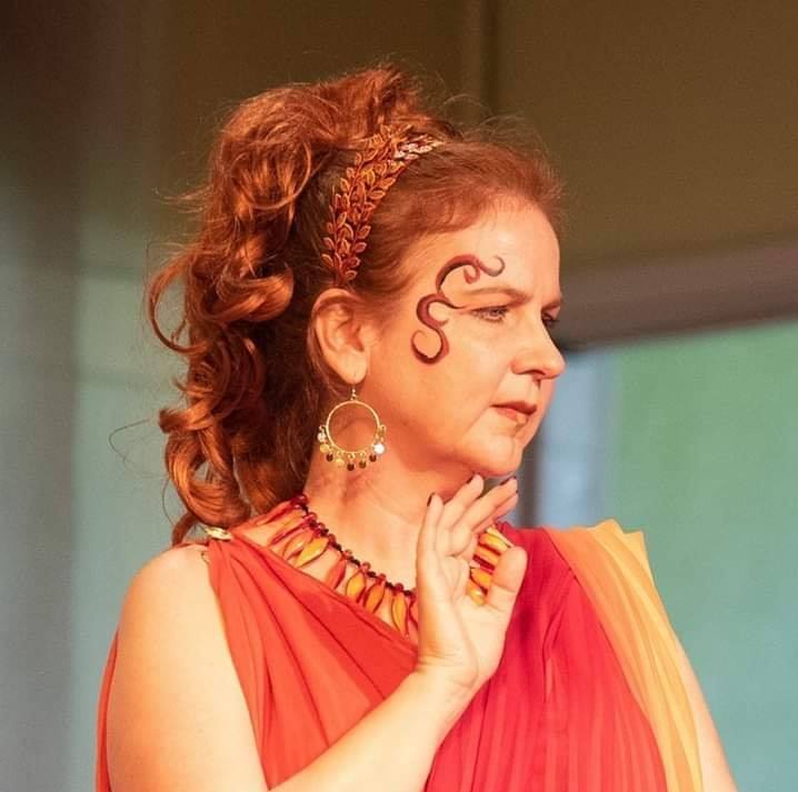 A woman wearing a red dress and earrings has a swirl painted on her face, representing Hestia.