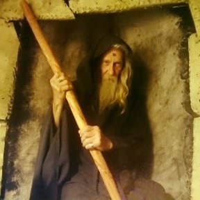 A man with a beard is holding a wooden stick, Terry Riley representing Charon.