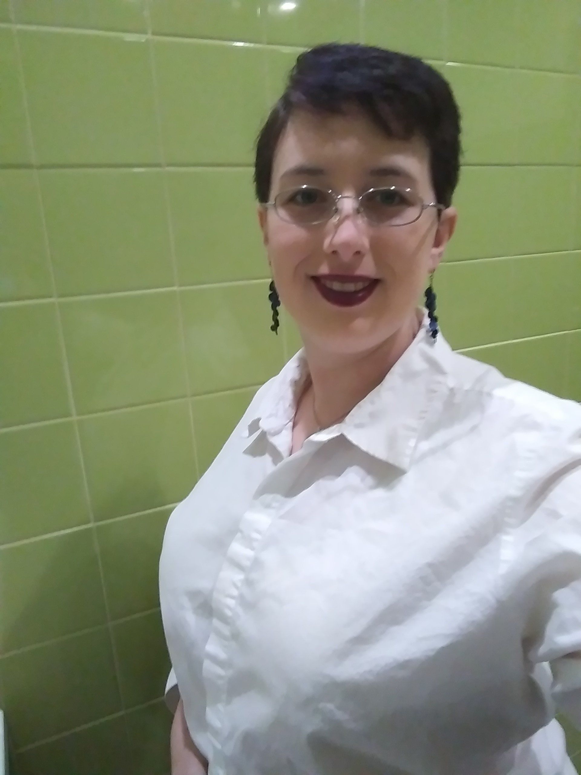 A woman wearing glasses and a white shirt is standing in front of a green tiled wall.