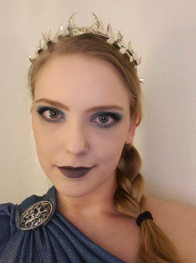 A woman wearing a crown and a blue dress is taking a selfie, representing Artemis.