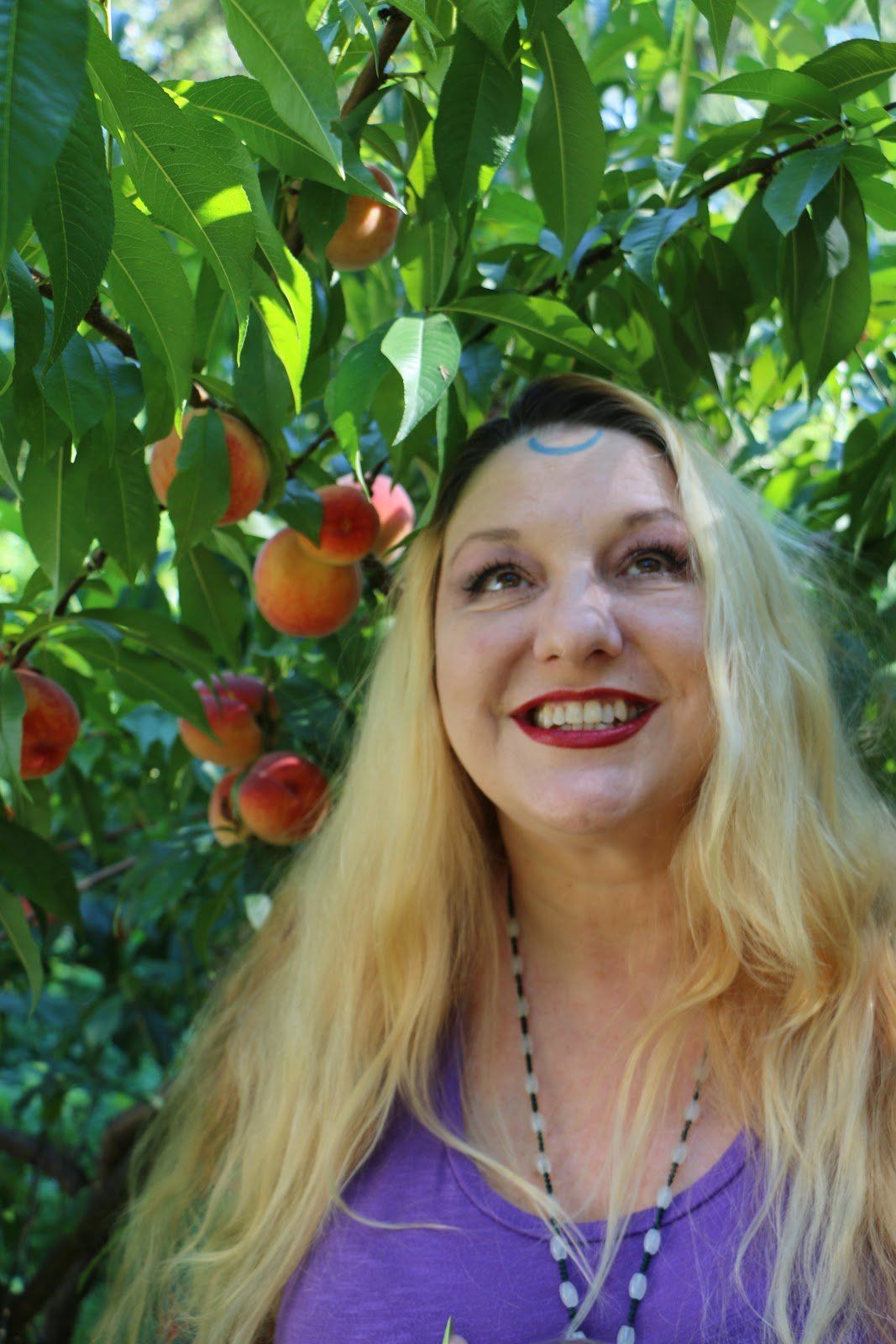 A woman is smiling in front of a peach tree, Belladonna LaVeau.