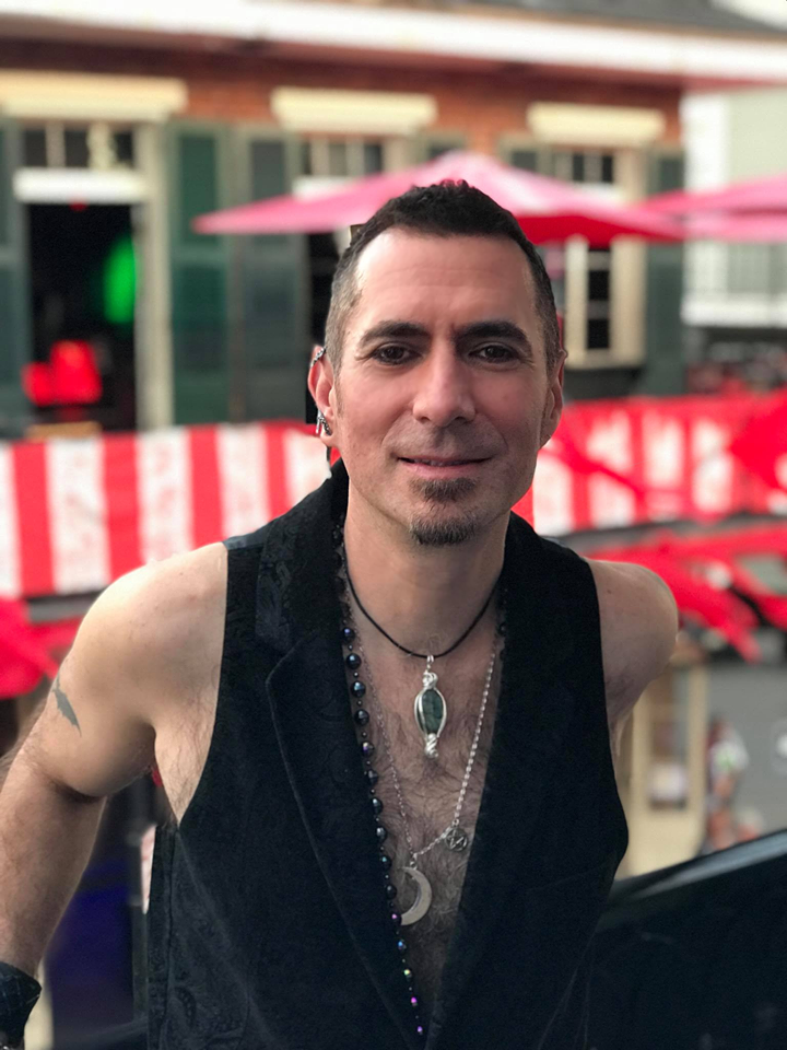 A man wearing a black vest and a necklace is standing in front of a building.