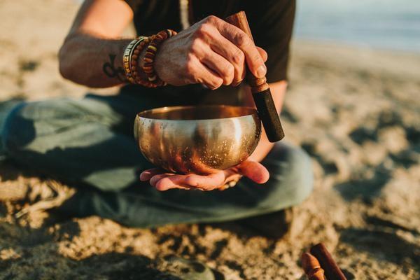 A man is sitting on the beach holding a singing bowl in his hands.