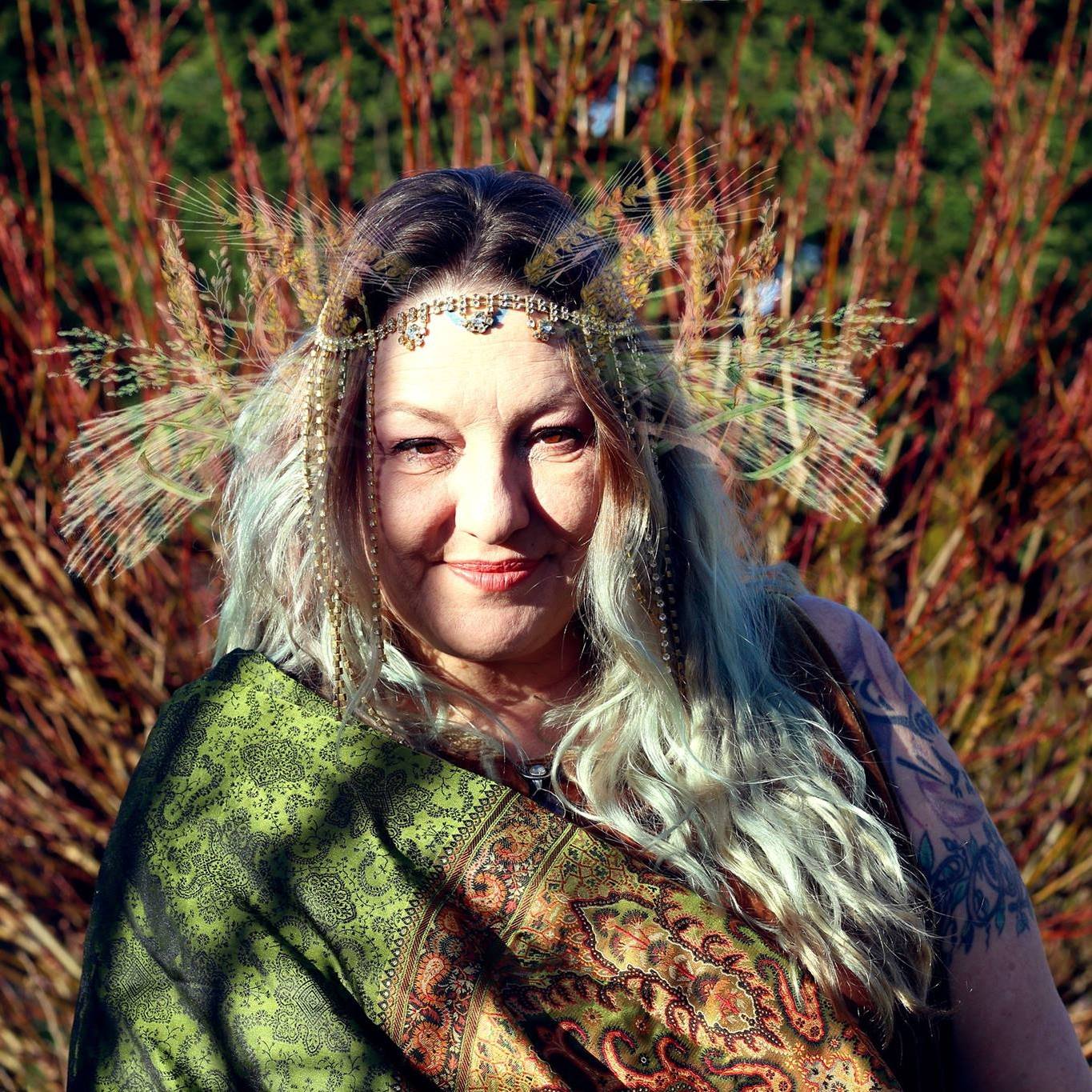 A woman wearing a headband and a scarf is standing in front of a bush, Belladonna LaVeau representing Demeter.