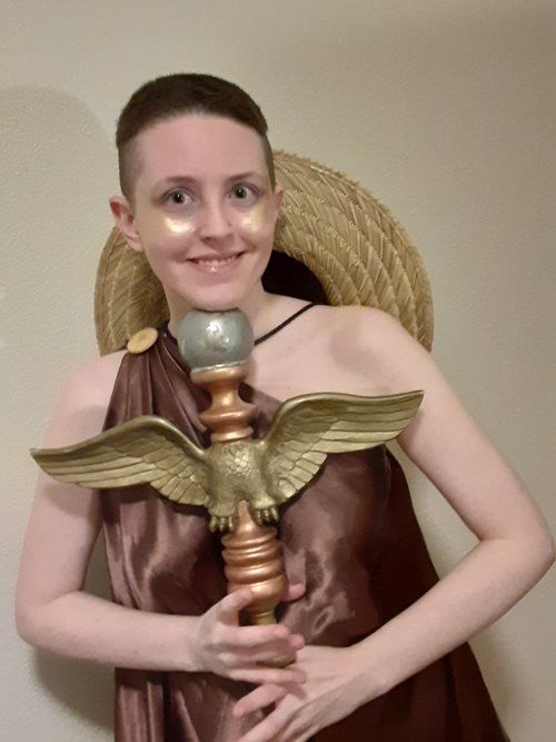 A woman in a brown dress is holding a statue of an eagle, Kel representing Hermes.