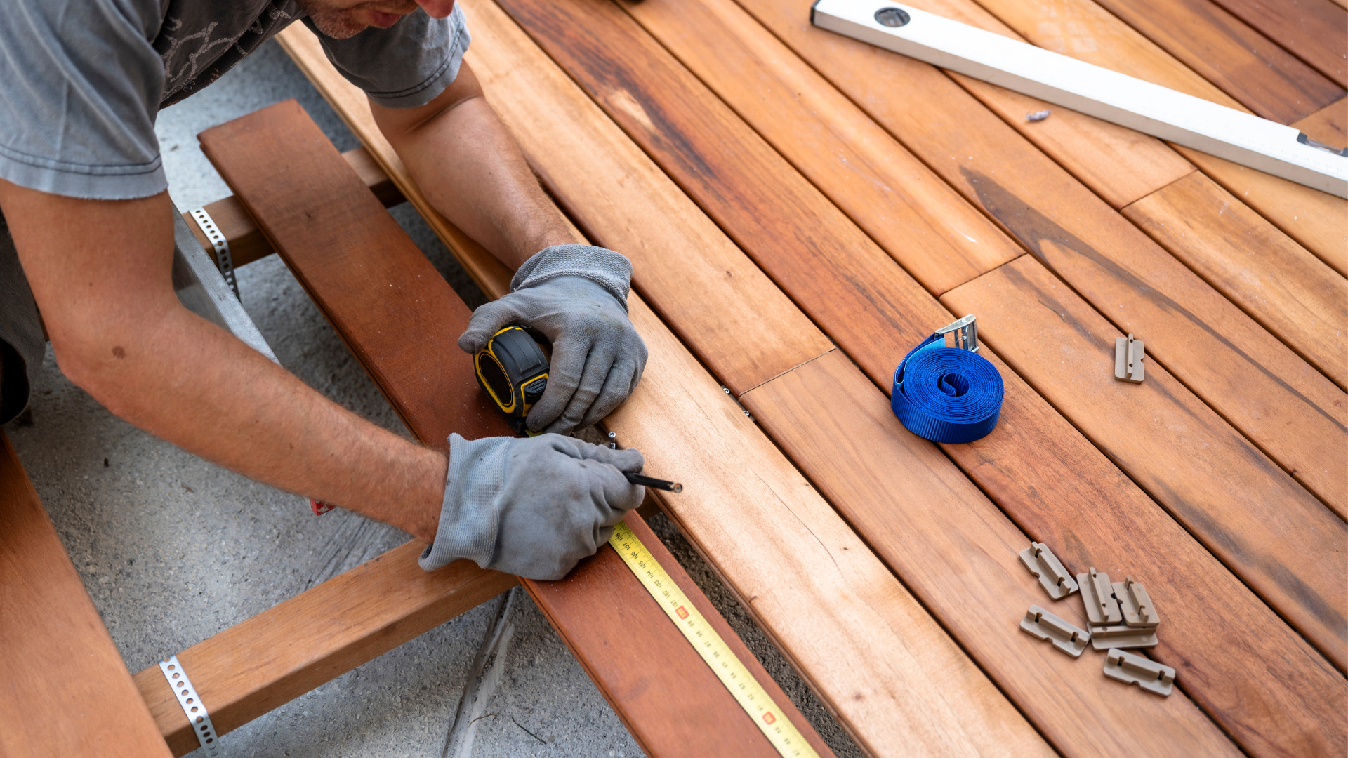 A person wearing work gloves measures and marks a wooden deck plank with a tape measure and pencil during construction.