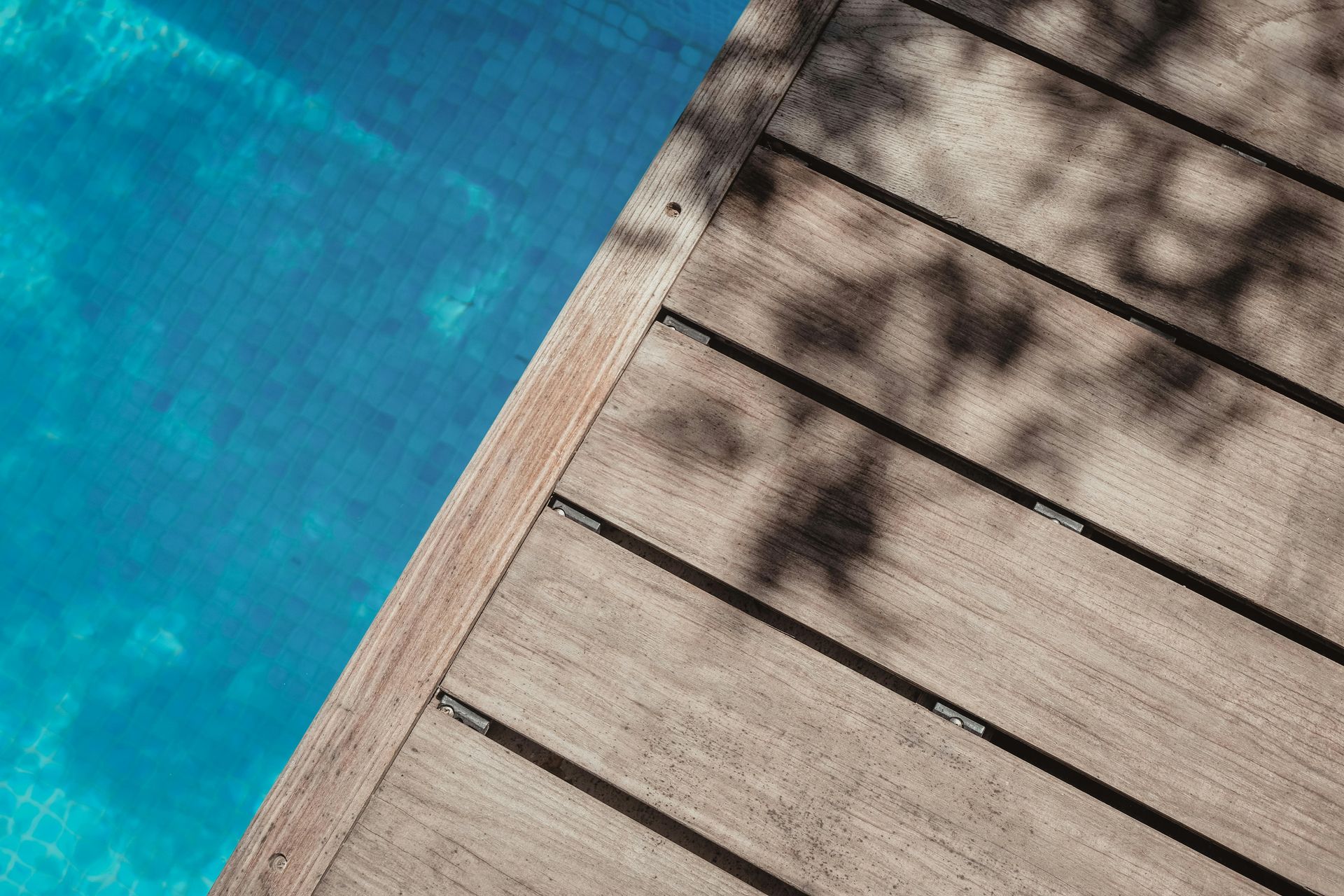 Wooden decking boards running diagonally beside a bright blue swimming pool, with leaf shadows cast on the wood.