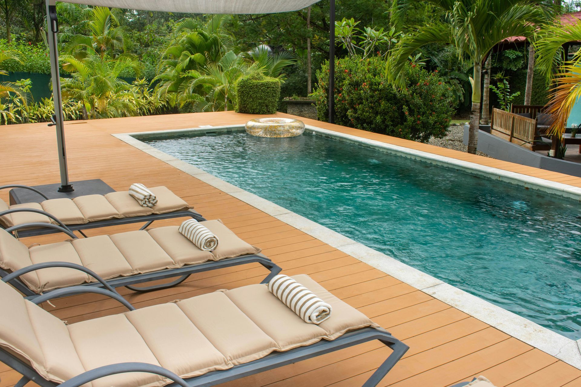 Three tan lounge chairs with striped pillows lined up on a wooden deck beside a rectangular outdoor swimming pool.