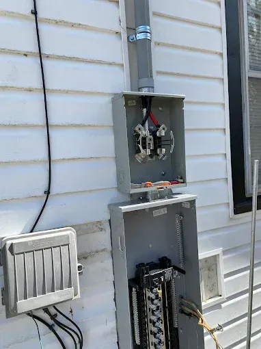 Electrical panel box on the side of a white house. Open box shows wires and circuits; grey conduit above.