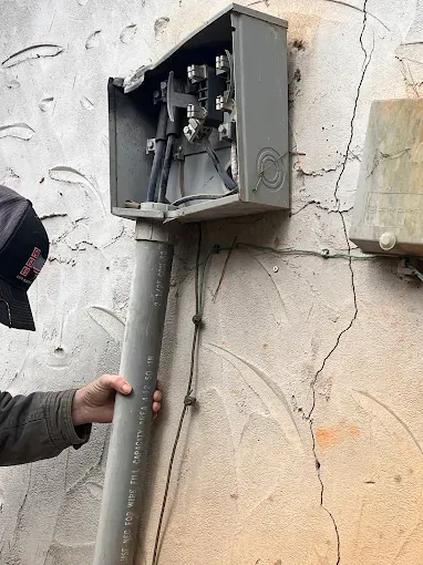 Damaged electrical box on a wall, conduit detached. A person holds the conduit.