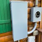 Gray electrical panel and outlet box on a wooden cabin wall, connected by conduit. Green shutter is visible.