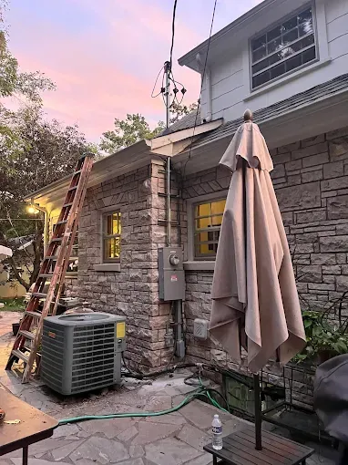 Exterior of a stone building with an air conditioner, ladder, closed umbrella, and pink sky.