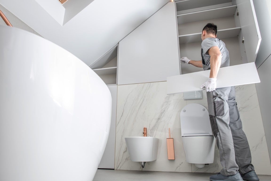 Worker installs an upper bathroom cabinet above a wall mounted toilet and sink in a modern bathroom.