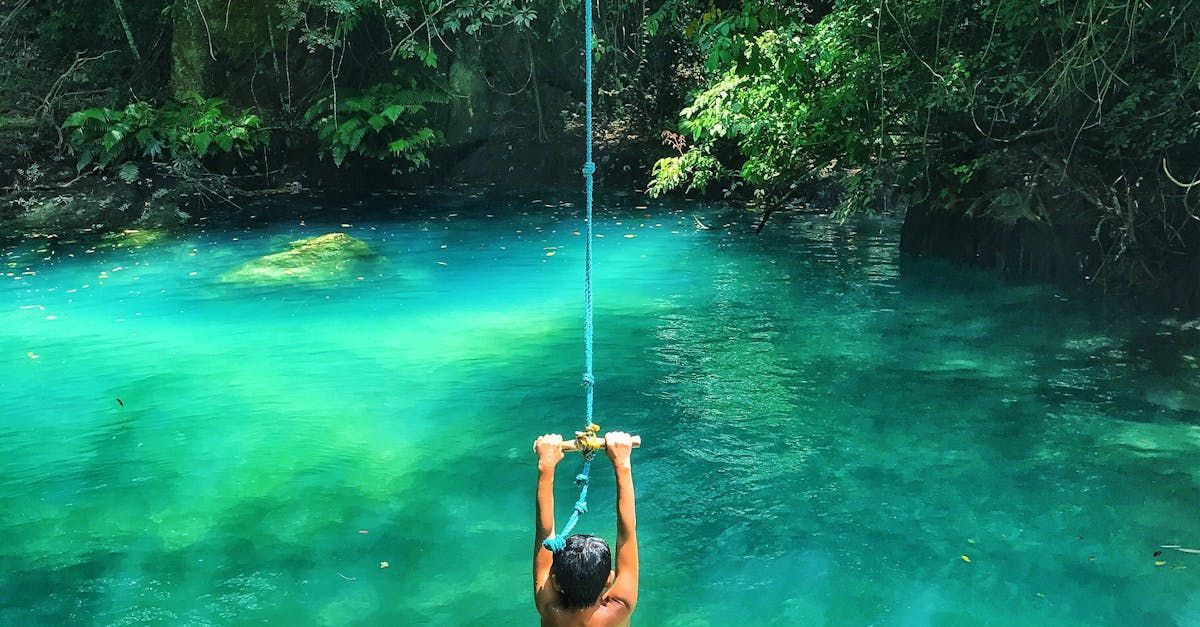 Person swinging on a rope into bright turquoise water. Surrounded by green trees.