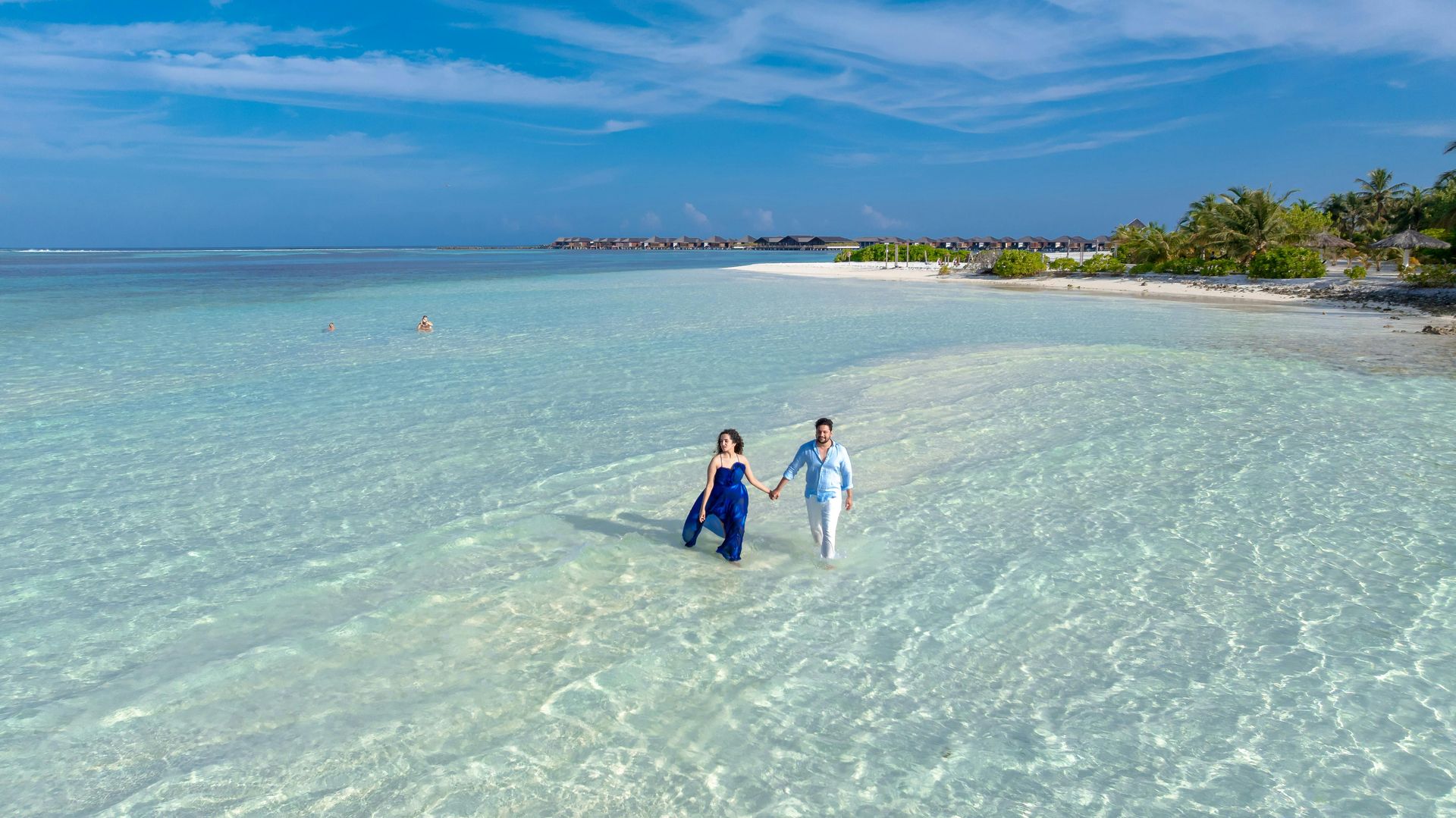 Couple walks hand-in-hand in shallow, clear turquoise water, with a sandy beach and blue sky in the background.