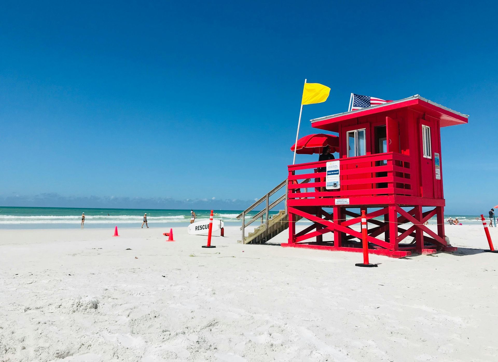 Bright red lifeguard station on a white sandy beach under a clear blue sky; yellow flag.