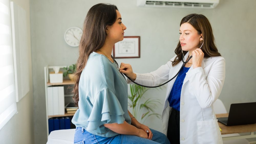 A clinician uses a stethoscope to examine a patient sitting on an exam table in a brightly lit medical office.