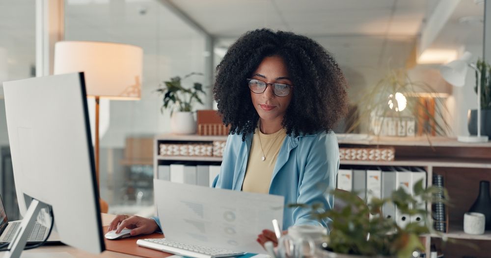 Woman with curly hair works at a computer, reviewing papers in a bright office.
